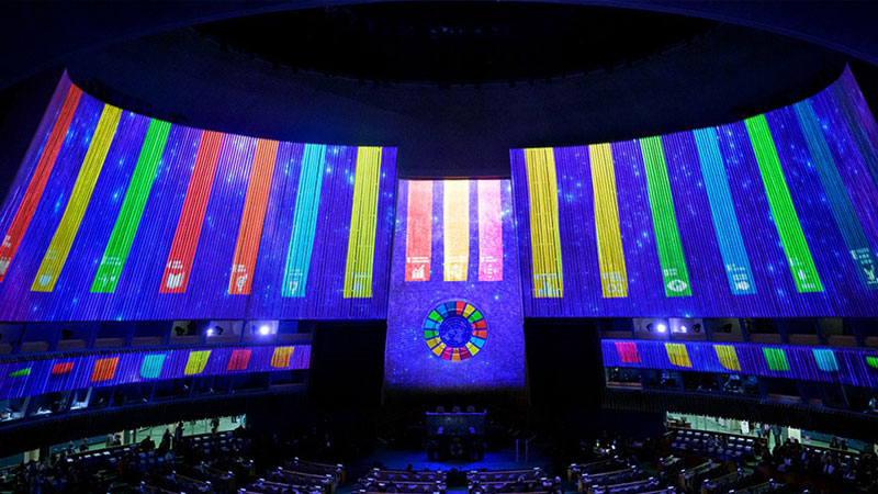 a photo no the UN Assembly Hall, with the SDGs wheel projected with light onto the walls and the in the UN emblem at the center